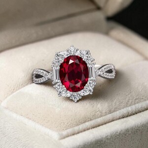 May include: A silver ring featuring a large, oval-cut, deep red gemstone surrounded by smaller, clear stones. The ring's band has an intricate, twisted design, and the setting is a halo style. The ring is displayed in a soft, off-white jewelry box.