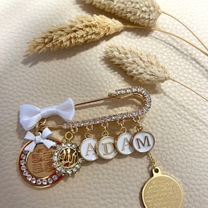 Könnte beinhalten: Goldfarbene Brosche mit dem Namen "ADAM" auf weißen Emaille-Kreisen. Die Brosche hat ein Sicherheitsnadel-Design, verziert mit klaren Kristallen und einer kleinen weißen Schleife. Zusätzliche goldene Charms und ein dekoratives Medaillon sind angebracht.