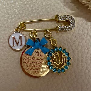 Op de afbeelding: Een goudkleurige veiligheidsspeld-broche met een met kristallen ingelegde kop, een witte ronde bedel met de letter M, een kleine blauwe strik, een ronde gouden hanger met Arabische kalligrafie en een bedel met blauwe kristallen rond het woord Allah.