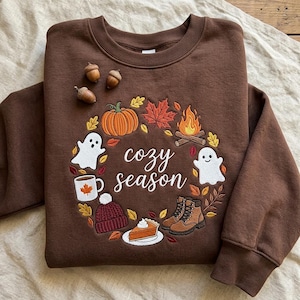 Peut inclure: Un sweat-shirt à col rond marron foncé avec une couronne brodée de motifs automnaux incluant une citrouille, un feu de camp, des fantômes, une tasse, un bonnet, des bottes et une part de tarte. Le texte "cozy season" est brodé en blanc.