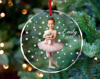 May include: A clear, round Christmas ornament with a photo of a young girl in a pink tutu holding a teddy bear. The ornament has the name "Emily" and the year "2025" printed on it, with snowflake accents and a red ribbon for hanging.