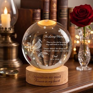 May include: A clear glass sphere memorial light on a round wooden base. The sphere features an etched cardinal bird, snowflakes, and the text: In Loving Memory, Those we love don't go away, they walk beside us every day. Unseen, unheard, but always near. Still loved, still missed, and very dear. Dad 1974-2026. The base reads: Always on my mind, Forever in my heart.
