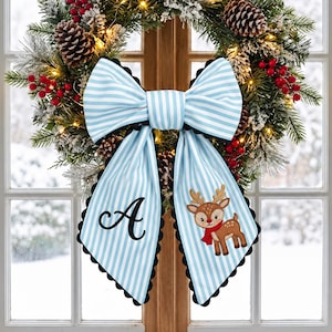 May include: A large decorative bow featuring light blue and white vertical stripes with a black scalloped edge. The left tail displays a black embroidered letter A, while the right tail features an embroidered brown reindeer wearing a red scarf.