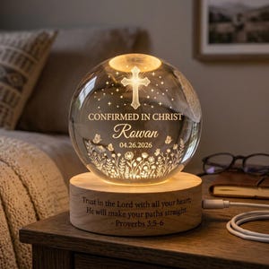 Könnte beinhalten: Ein Nachtlicht in Form einer klaren Glaskugel auf einem runden Holzsockel. Die Kugel zeigt ein eingraviertes Kreuz, Sterne, Schmetterlinge, Blumen und den Text "CONFIRMED IN CHRIST Rowan 04.26.2026". Der Holzsockel ist mit "Trust in the Lord with all your heart; He will make your paths straight. - Proverbs 3:5-6" graviert.