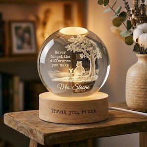 Può includere: Una luce notturna a sfera in vetro trasparente con un design inciso al laser raffigurante un orso e un piccolo animale sotto un albero. Il testo recita "Never forget the difference you make" e "Mrs. Sloane". La base rotonda in legno riporta l'incisione "Thank you, Frank".