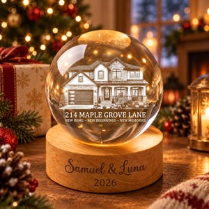 May include: A clear glass sphere on a round wooden base. The sphere features an etched house illustration with the text 214 MAPLE GROVE LANE NEW HOME NEW BEGINNINGS NEW MEMORIES. The wooden base is engraved with the names Samuel & Luna and the year 2026.
