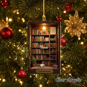 May include: A rectangular hanging ornament featuring a miniature library scene with multiple shelves filled with colorful books, a rolling ladder, a small potted plant, and a wall lamp. The text reads To plant a garden is to believe in tomorrow.