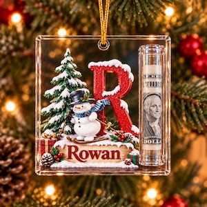 May include: A clear acrylic Christmas ornament featuring a red glittery letter R, a snowman on skis, a snow-covered pine tree, and a cylindrical tube containing a rolled one-dollar bill. The base displays the name Rowan in red lettering.