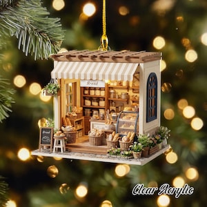 May include: A miniature bakery ornament featuring a beige striped awning, a wooden pergola roof, and a sign labeled Bread. The interior displays shelves with jars, baskets of bread, a glass display case, and a small chalkboard sign labeled Bread.