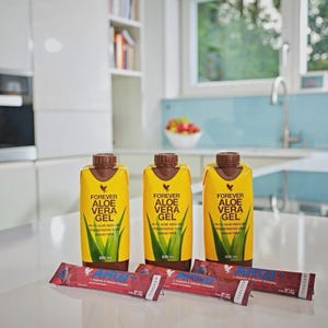 Op de afbeelding: Drie gele en bruine Aloe Vera Gel flessen en drie rode ARGI+ zakjes worden getoond op een wit aanrechtblad. De flessen hebben een bruine dop en de tekst "FOREVER ALOE VERA GEL" met een groene aloë vera plant afbeelding. De zakjes lezen "ARGI+ L-Arginine & Vitamin Complex".