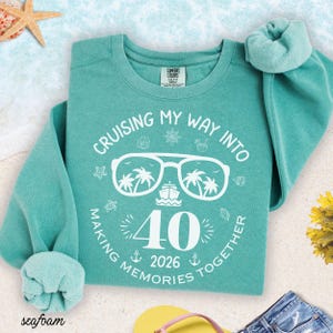 May include: Seafoam green sweatshirt with white graphic text and illustrations. The text reads "Cruising My Way Into Making Memories Together" with "40 2026" and a sunglasses design with palm trees and a cruise ship. The sweatshirt has a crew neck and long sleeves.