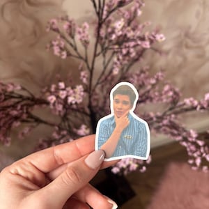 May include: A sticker featuring a young man in a blue striped shirt, with his hand on his chin. The sticker is held in front of a pink floral arrangement. The background has a soft, blurred pattern.