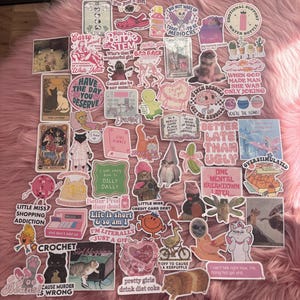 Pode incluir: Uma coleção de autocolantes coloridos com vários designs e texto. Os autocolantes apresentam ilustrações de gatos, personagens de desenhos animados e frases como "I'm just a girl" e "Better late than ugly". Os autocolantes estão dispostos numa superfície rosa e fofa.