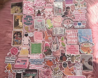 May include: A collection of colorful stickers with various designs and text. The stickers feature illustrations of cats, cartoon characters, and phrases like "I'm just a girl" and "Better late than ugly." The stickers are arranged on a pink, fluffy surface.