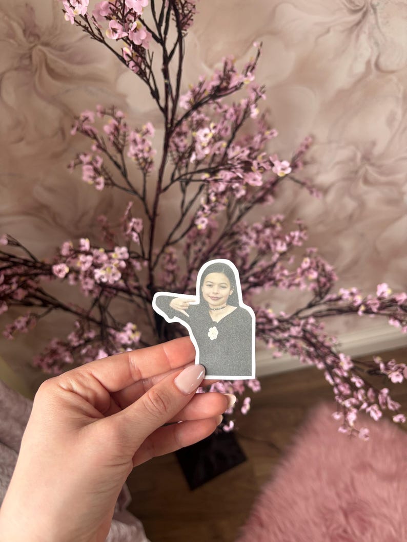 May include: A sticker featuring a person in a black top with a necklace, held in a hand. The background includes a pink floral arrangement and a pink rug.