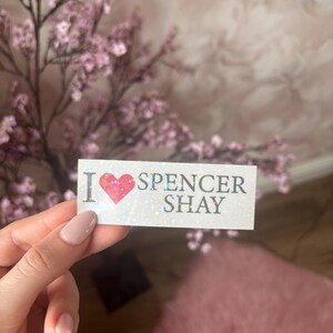 May include: A white sticker with the text "I ❤️ SPENCER SHAY" in a serif font. The heart is pink. The sticker is held in front of a blurred background of pink flowers and a pink rug.