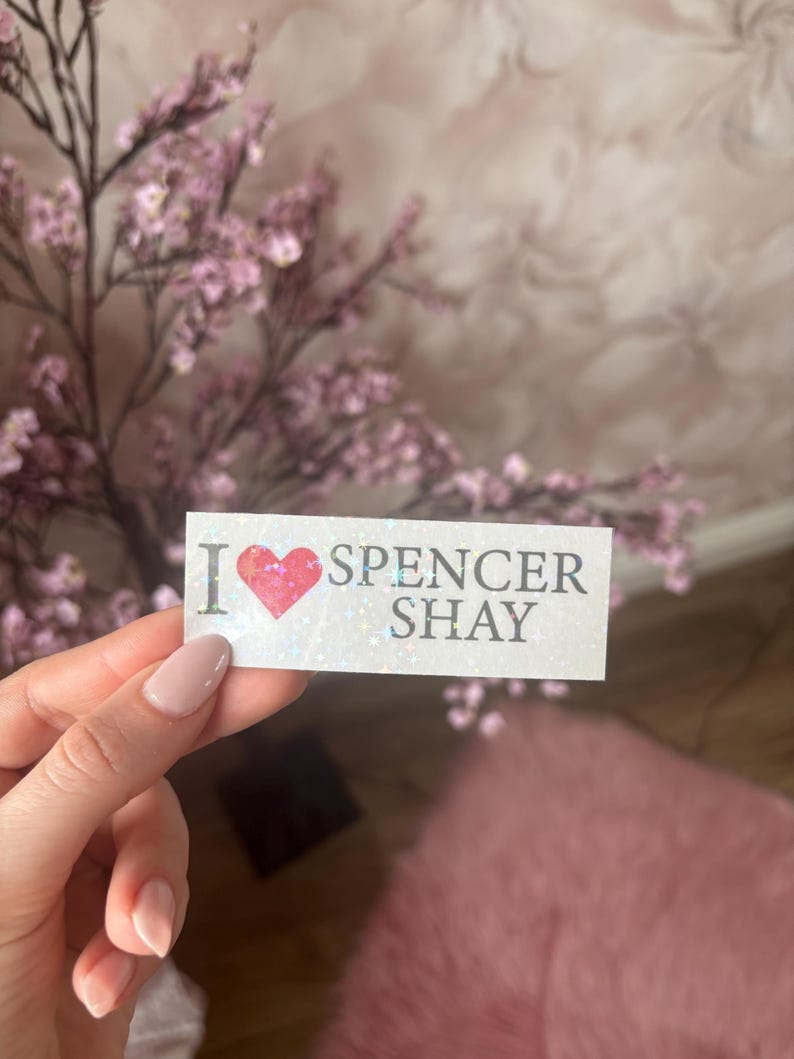 May include: A white sticker with the text "I ❤️ SPENCER SHAY" in a serif font. The heart is pink. The sticker is held in front of a blurred background of pink flowers and a pink rug.