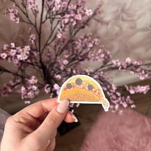 May include: A die-cut sticker shaped like a taco with a yellow shell, red sauce, and three dark gray meatballs. The sticker features a white border and a cartoon-style illustration of the food item with a small tail of sauce at the bottom.