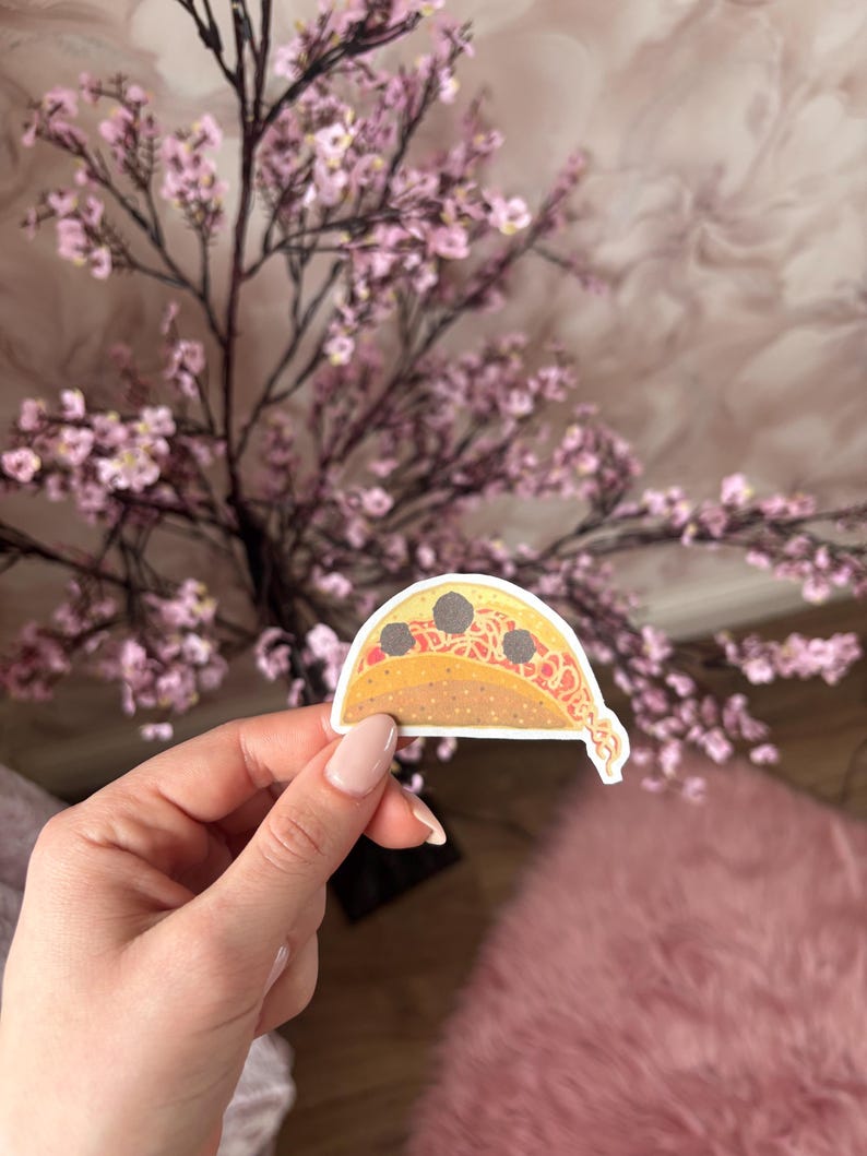 May include: A die-cut sticker shaped like a taco with a yellow shell, red sauce, and three dark gray meatballs. The sticker features a white border and a cartoon-style illustration of the food item with a small tail of sauce at the bottom.