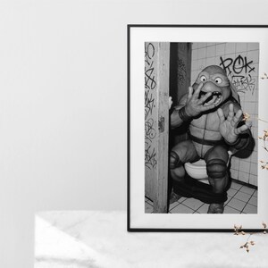 May include: Monochrome framed print depicting a Teenage Mutant Ninja Turtle character in a bathroom. The character is seated on a toilet, with a startled expression. Graffiti art is visible on the walls.