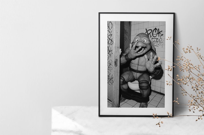 May include: Monochrome framed print depicting a Teenage Mutant Ninja Turtle character in a bathroom. The character is seated on a toilet, with a startled expression. Graffiti art is visible on the walls.