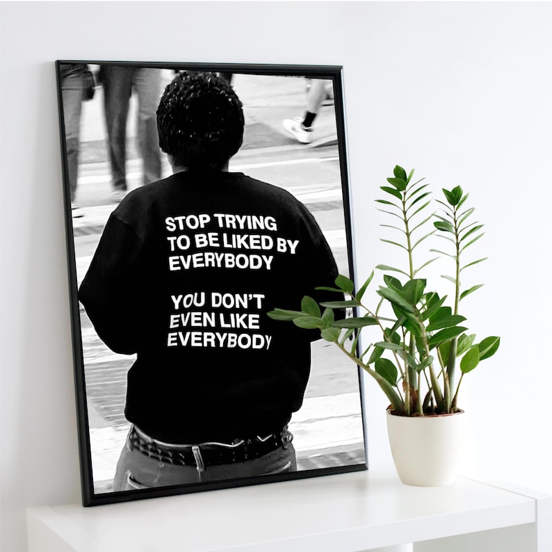 K&ouml;nnte beinhalten: Schwarz-Wei&szlig;-Druck in einem Rahmen, der eine Person in einem schwarzen Oberteil mit dem Text "STOP TRYING TO BE LIKED BY EVERYBODY YOU DON'T EVEN LIKE EVERYBODY" zeigt. Eine kleine Topfpflanze steht auf einer wei&szlig;en Oberfl&auml;che.