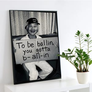 Puede incluir: Impresión en blanco y negro enmarcada que muestra a una persona sonriente con una gorra con la palabra "money". La persona sostiene un letrero con el texto "To be ballin, you gotta b-all-in". La impresión está en un estante blanco.