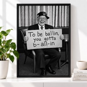 Pode incluir: Uma fotografia emoldurada a preto e branco de uma pessoa mais velha com boné, óculos e fato, sentada numa cadeira e segurando um cartaz com o texto: To be ballin, you gotta b-all-in. A imagem é apresentada numa moldura preta simples.