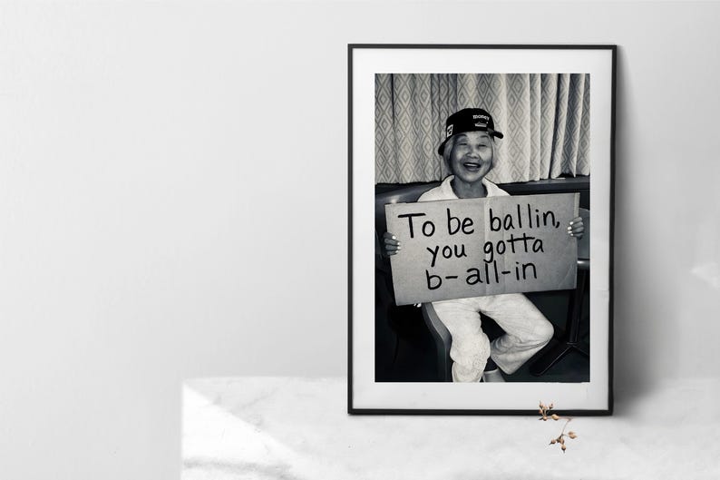 May include: Black and white framed print featuring a person wearing a baseball cap and holding a sign. The sign reads "To be ballin, you gotta b-all-in". The person is smiling.