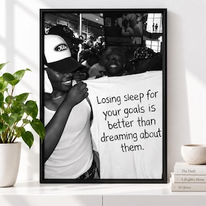 May include: A black and white framed print featuring a person wearing a white tank top and a baseball cap, holding up a white t-shirt with the text: Losing sleep for your goals is better than dreaming about them. The background shows a crowd.
