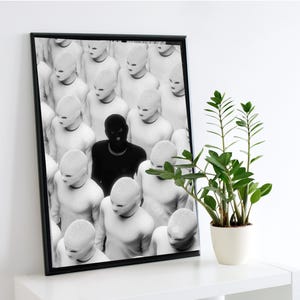 Puede incluir: Impresión artística enmarcada en blanco y negro que presenta una multitud de figuras vestidas de blanco y enmascaradas, con una figura en negro. La obra de arte se exhibe en una estantería blanca.