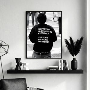 K&ouml;nnte beinhalten: Schwarz-Wei&szlig;-Druck in einem Rahmen, der eine Person in einem schwarzen Sweatshirt mit dem Text "STOP TRYING TO BE LIKED BY EVERYBODY YOU DON'T EVEN LIKE EVERYBODY" zeigt. Das Bild ist eine Fotografie einer Person in einer Menschenmenge.