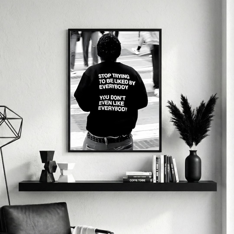 K&ouml;nnte beinhalten: Schwarz-Wei&szlig;-Druck in einem Rahmen, der eine Person in einem schwarzen Sweatshirt mit dem Text "STOP TRYING TO BE LIKED BY EVERYBODY YOU DON'T EVEN LIKE EVERYBODY" zeigt. Das Bild ist eine Fotografie einer Person in einer Menschenmenge.