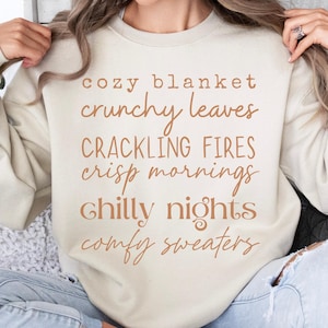 May include: A beige crewneck sweatshirt featuring brown text that reads: cozy blanket, crunchy leaves, CRACKLING FIRES, crisp mornings, chilly nights, comfy sweaters. The garment has long sleeves and a ribbed neckline, cuffs, and hem.