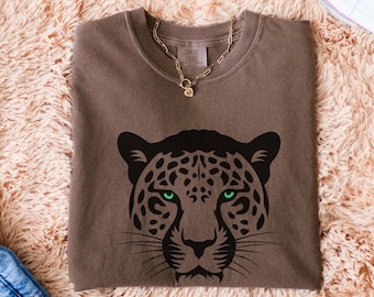 May include: A brown t-shirt featuring a black leopard face design with green eyes. The shirt is paired with a gold necklace, blue jeans, and white sneakers. The shirt is folded on a fluffy, peach-colored rug.