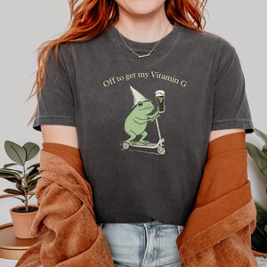 May include: Dark gray t-shirt with the text "Off to get my Vitamin G." The shirt features a cartoon frog wearing a wizard hat, riding a scooter, and holding a beer. The frog is green, and the beer is in a glass.