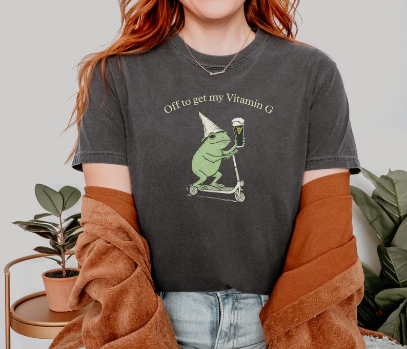 May include: Dark gray t-shirt with the text "Off to get my Vitamin G." The shirt features a cartoon frog wearing a wizard hat, riding a scooter, and holding a beer. The frog is green, and the beer is in a glass.