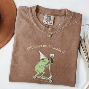 May include: Brown Comfort Colors t-shirt with the phrase "Off to get my Vitamin G" and a graphic of a green frog riding a scooter and holding a beer. A silver necklace and a brown hat are also in the image.