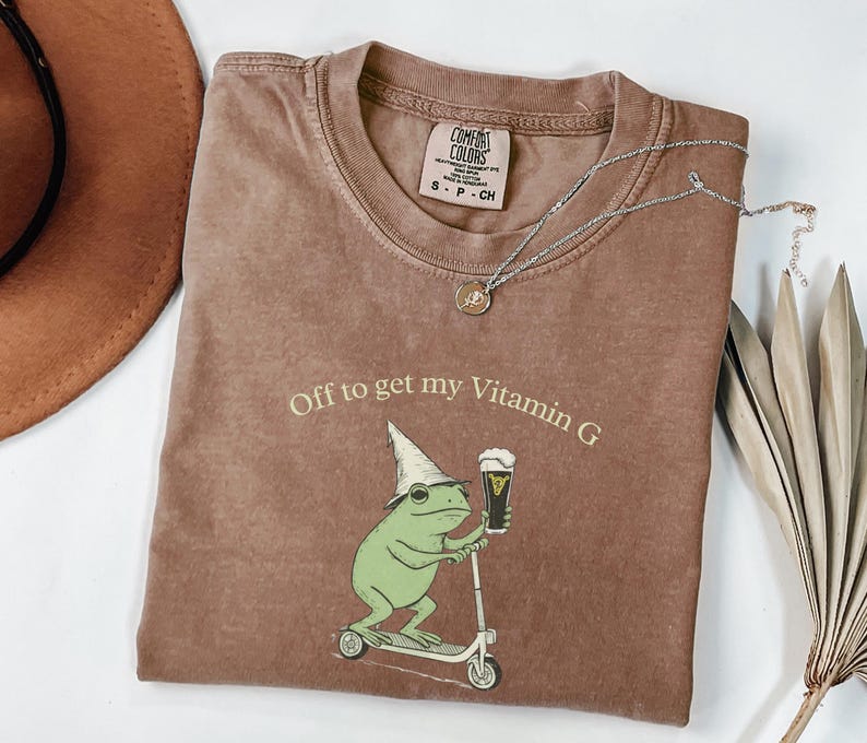 May include: Brown Comfort Colors t-shirt with the phrase "Off to get my Vitamin G" and a graphic of a green frog riding a scooter and holding a beer. A silver necklace and a brown hat are also in the image.