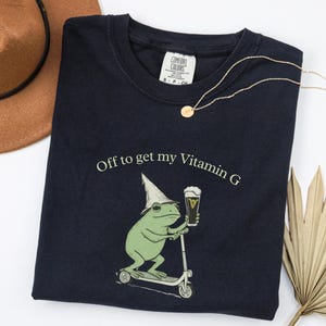May include: Navy blue t-shirt with the text "Off to get my Vitamin G" and a graphic of a green frog riding a scooter and holding a beer. A gold necklace and a brown hat are also in the image.