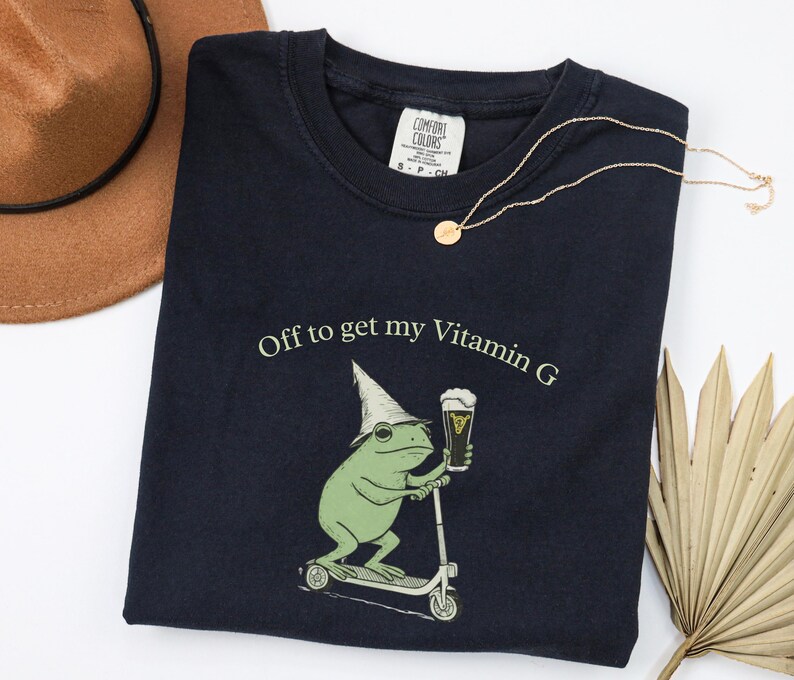 May include: Navy blue t-shirt with the text "Off to get my Vitamin G" and a graphic of a green frog riding a scooter and holding a beer. A gold necklace and a brown hat are also in the image.
