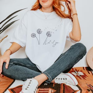 May include: White t-shirt with a dandelion graphic and the words "Let it go" in a script font. The shirt is paired with blue jeans and white sneakers. A straw hat and a woven basket are also visible.
