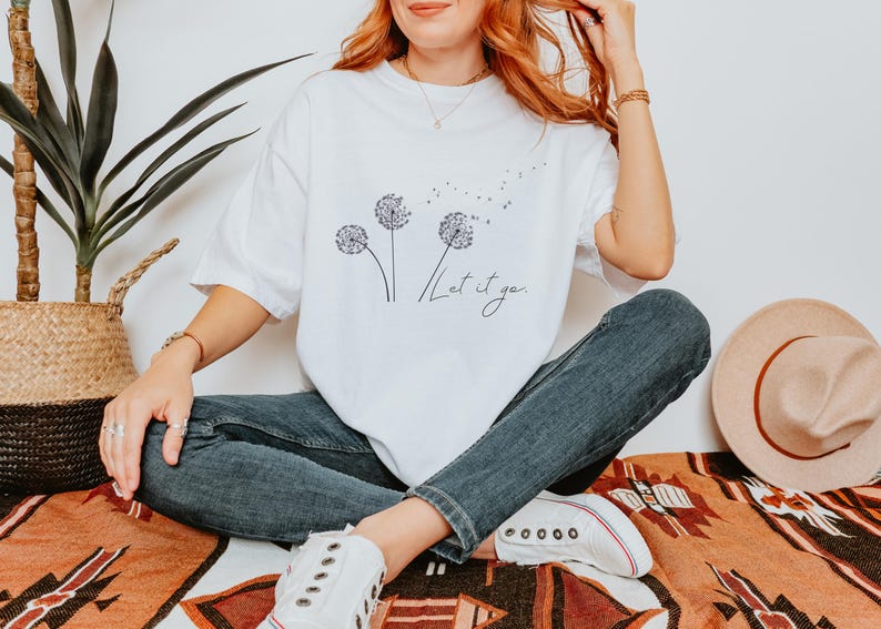 May include: White t-shirt with a dandelion graphic and the words "Let it go" in a script font. The shirt is paired with blue jeans and white sneakers. A straw hat and a woven basket are also visible.