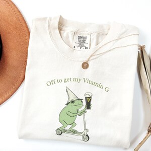May include: Cream-colored t-shirt with a graphic of a green frog riding a scooter and holding a pint of beer. The text "Off to get my Vitamin G" is printed above the frog. A gold necklace and a brown hat are also visible.