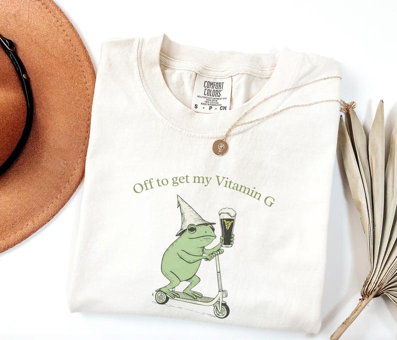 May include: Cream-colored t-shirt with a graphic of a green frog riding a scooter and holding a pint of beer. The text "Off to get my Vitamin G" is printed above the frog. A gold necklace and a brown hat are also visible.