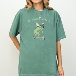 May include: A green t-shirt with the text "Off to get my Vitamin G." The shirt features a cartoon frog riding a scooter and holding a beer. The frog wears a wizard hat. The shirt is a solid color.