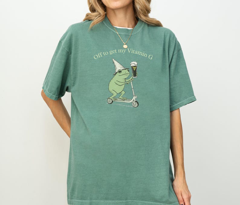 May include: A green t-shirt with the text "Off to get my Vitamin G." The shirt features a cartoon frog riding a scooter and holding a beer. The frog wears a wizard hat. The shirt is a solid color.