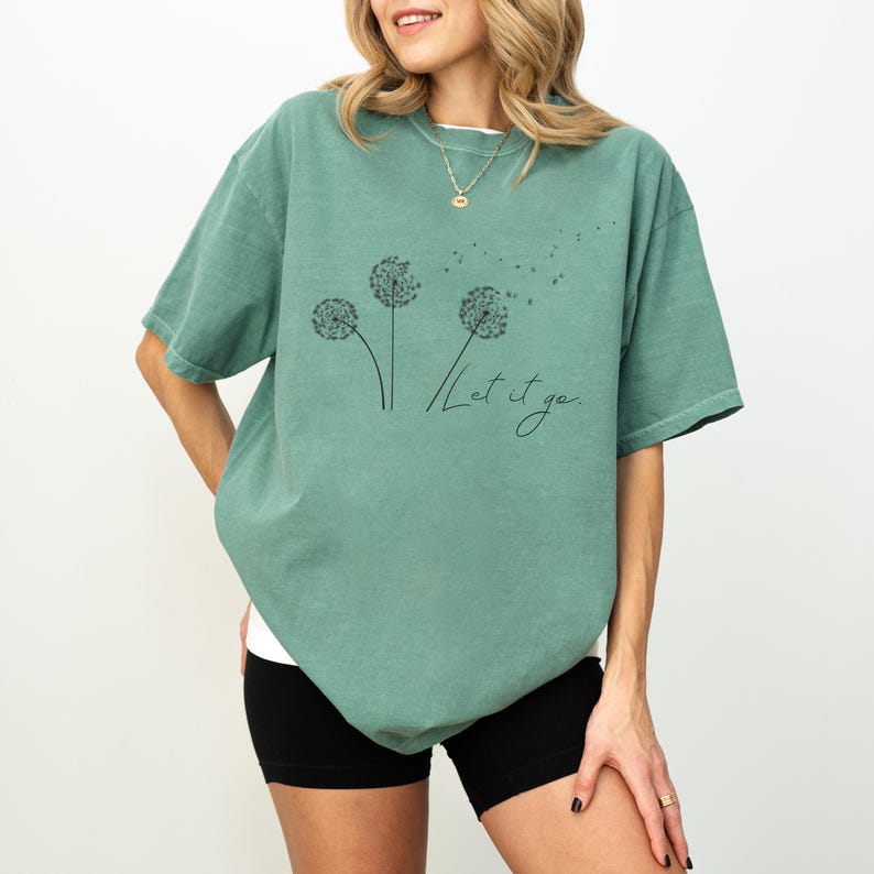 May include: Sage green t-shirt featuring a dandelion graphic with the words "Let it go" in a handwritten font. The shirt has short sleeves and a relaxed fit, suitable for casual wear.