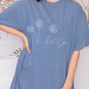 May include: A blue t-shirt featuring a dandelion design with the words "Let it go" in a cursive font. The shirt has a relaxed fit and short sleeves. The dandelion design is white and delicate.