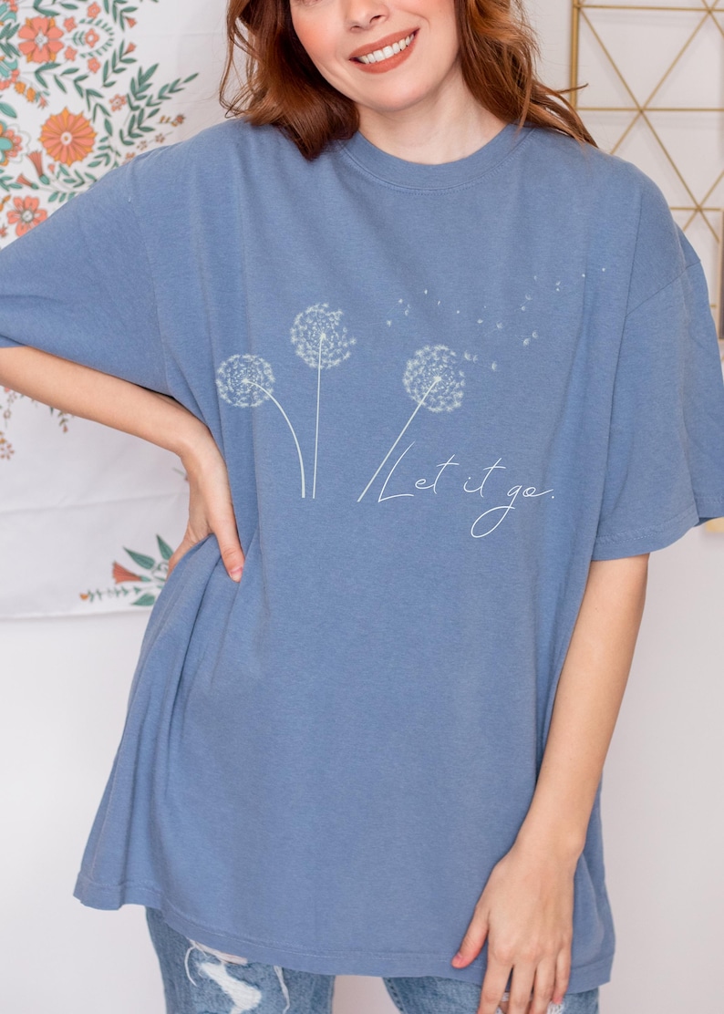 May include: A blue t-shirt featuring a dandelion design with the words "Let it go" in a cursive font. The shirt has a relaxed fit and short sleeves. The dandelion design is white and delicate.